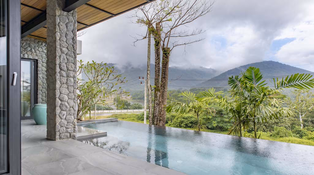 A three-bedroom villa in Thalang at dusk, with a mountain view and an illuminated swimming pool.