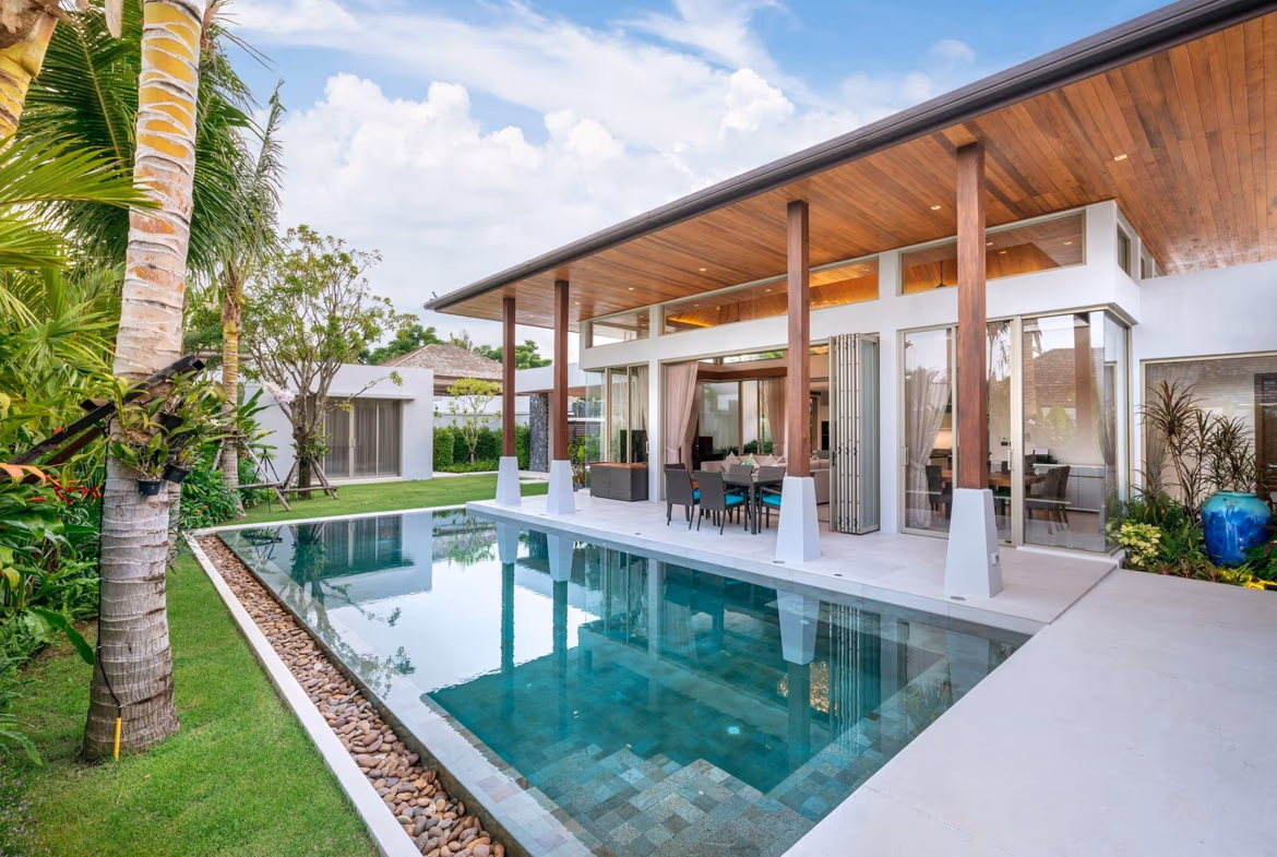 A view of a modern swimming pool and a patio area, with a wooded hillside in the background.
