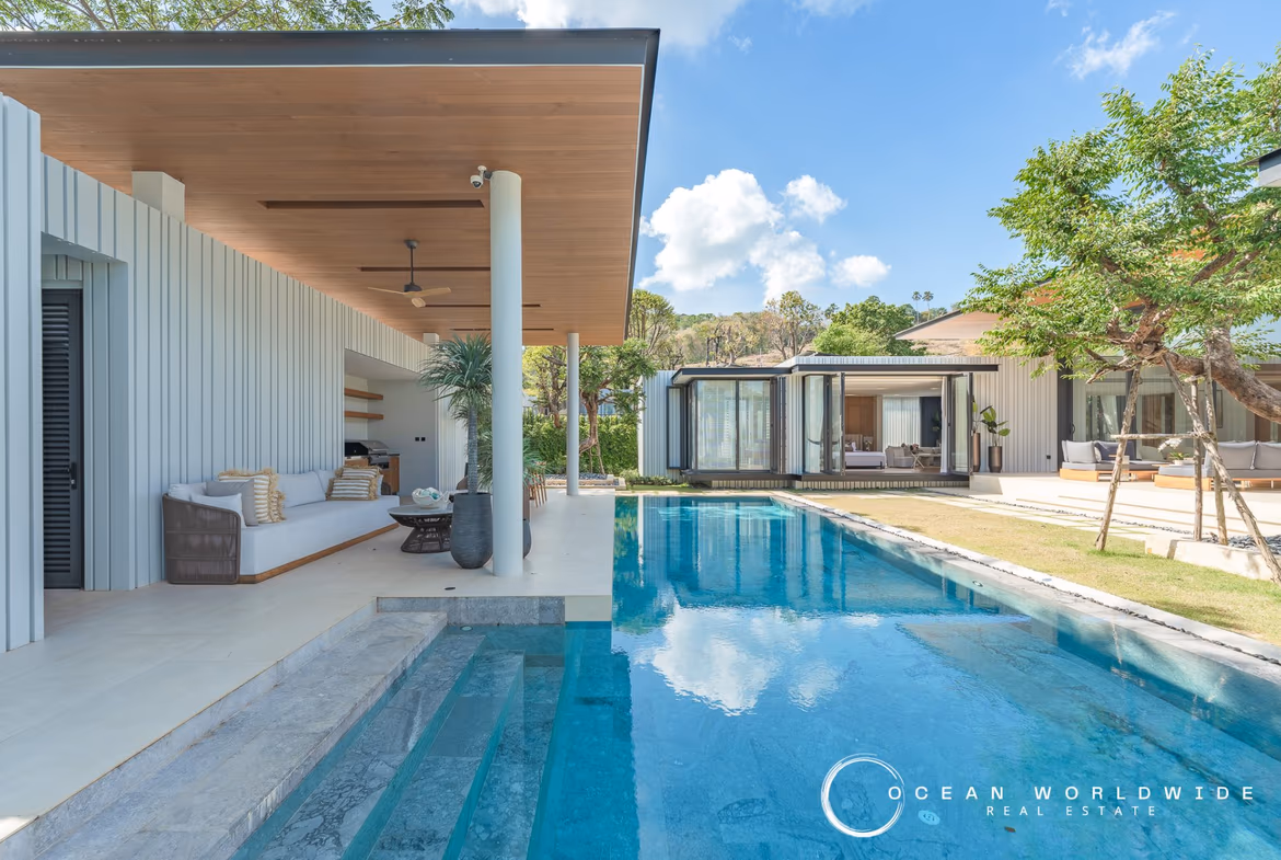 A spacious living room with large windows looks out onto a private swimming pool and a lush tropical landscape.