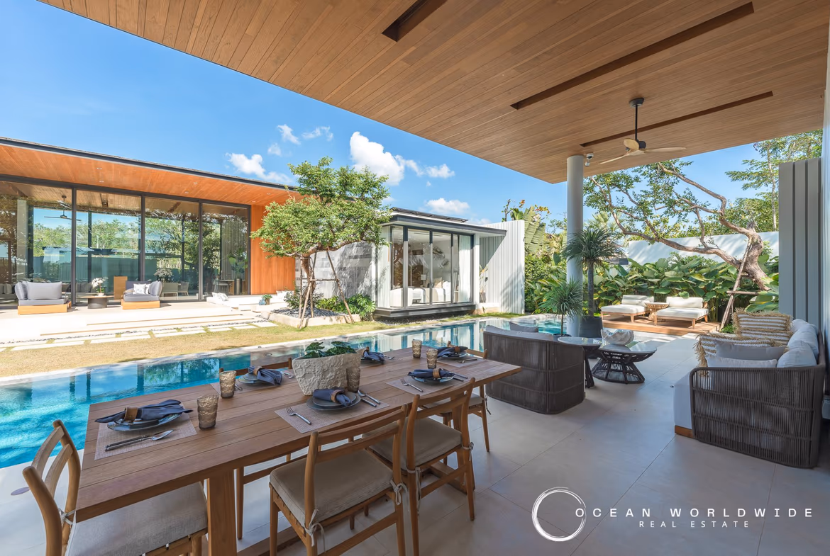 A luxurious living room with a high ceiling, large windows looking out to a garden and swimming pool, and a staircase leading to a second floor.
