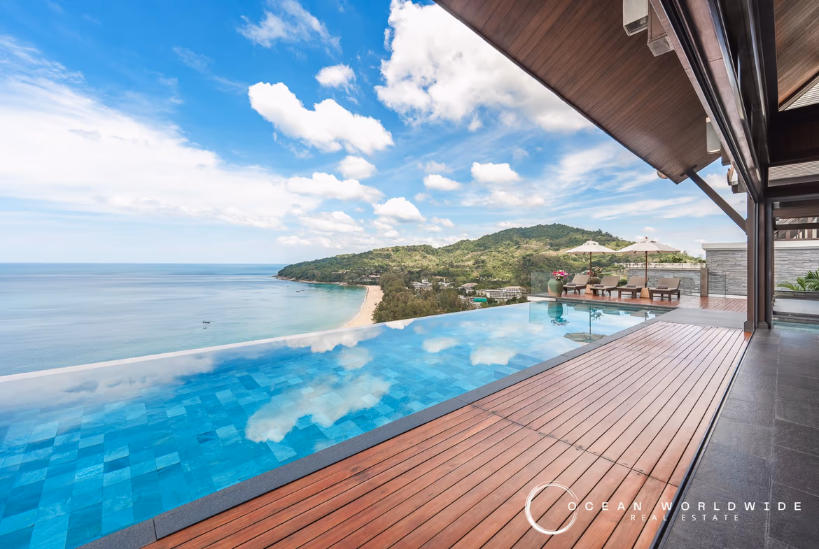 A luxurious, five-bedroom villa with a white exterior, multiple balconies, and a red tile roof overlooks a lush, green landscape in Bang Tao. A swimming pool is visible in the foreground.