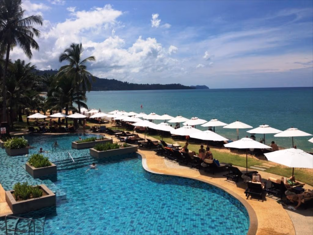 A modern hotel room features a large bed, light wood floors, and a private balcony overlooking a pool and palm trees.