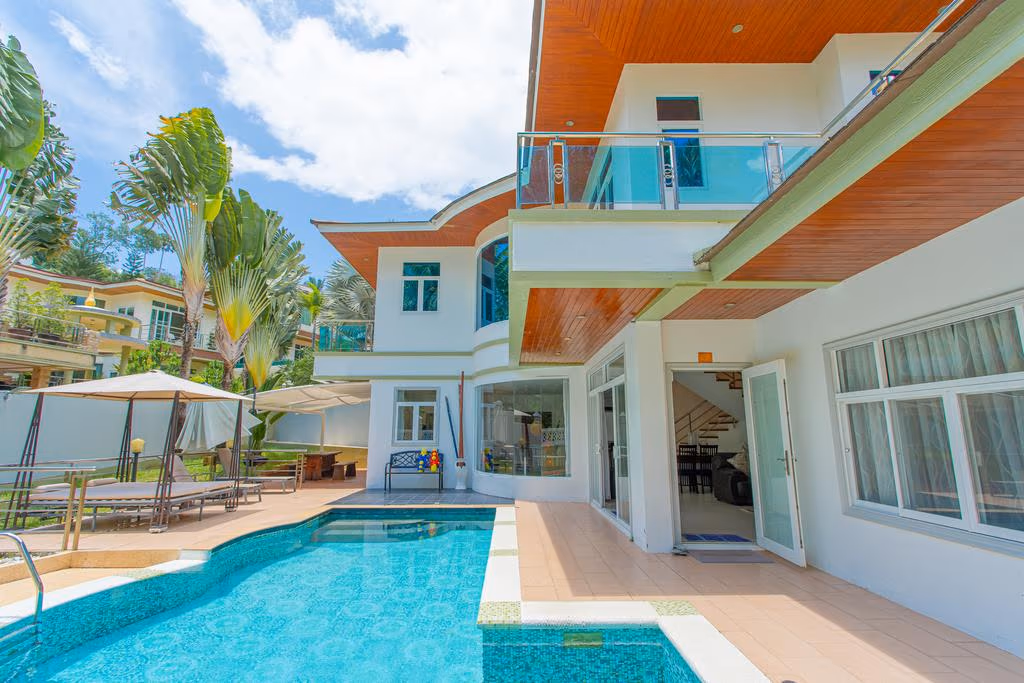 A four-bedroom villa in Karon with white walls and a red-tile roof. It has a spacious exterior with a swimming pool and surrounded by lush palm trees.