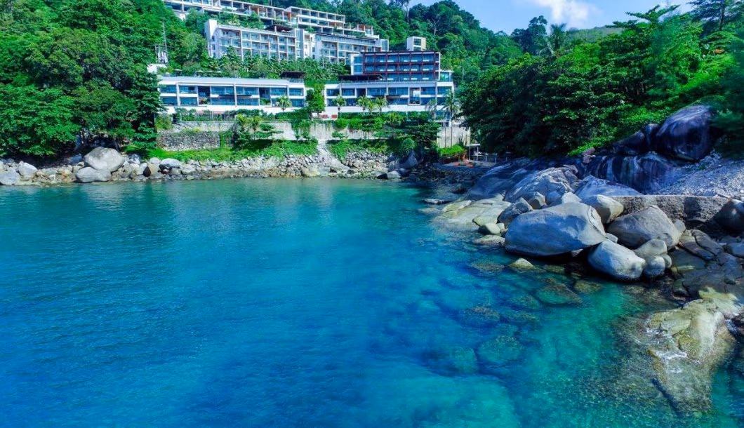 A modern beachfront hotel with a multi-story, white facade directly facing the ocean in Kamala. Palm trees line the entrance and part of the building.