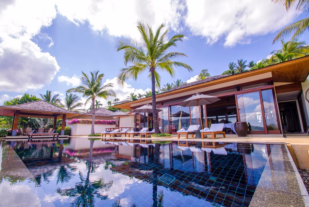 This 4-bedroom villa in Cherngtalay features a modern exterior and sea views. The multi-story white building is surrounded by lush tropical landscaping under a clear sky.
