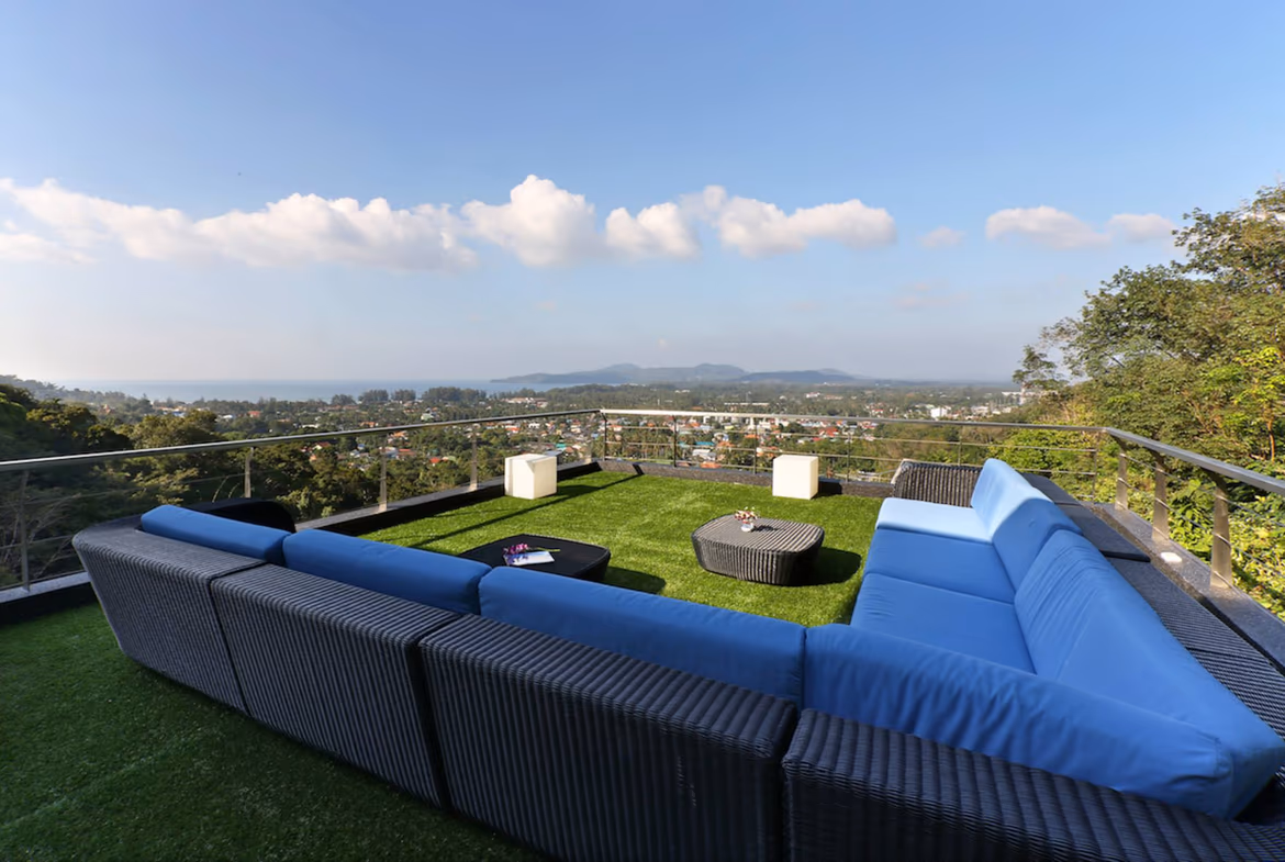 Several white buildings are situated at different levels on a lush green hillside, with the sea visible in the background and a large pool in the foreground.