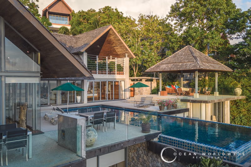 An elevated view from a balcony or patio shows a swimming pool and the ocean beyond tropical trees.