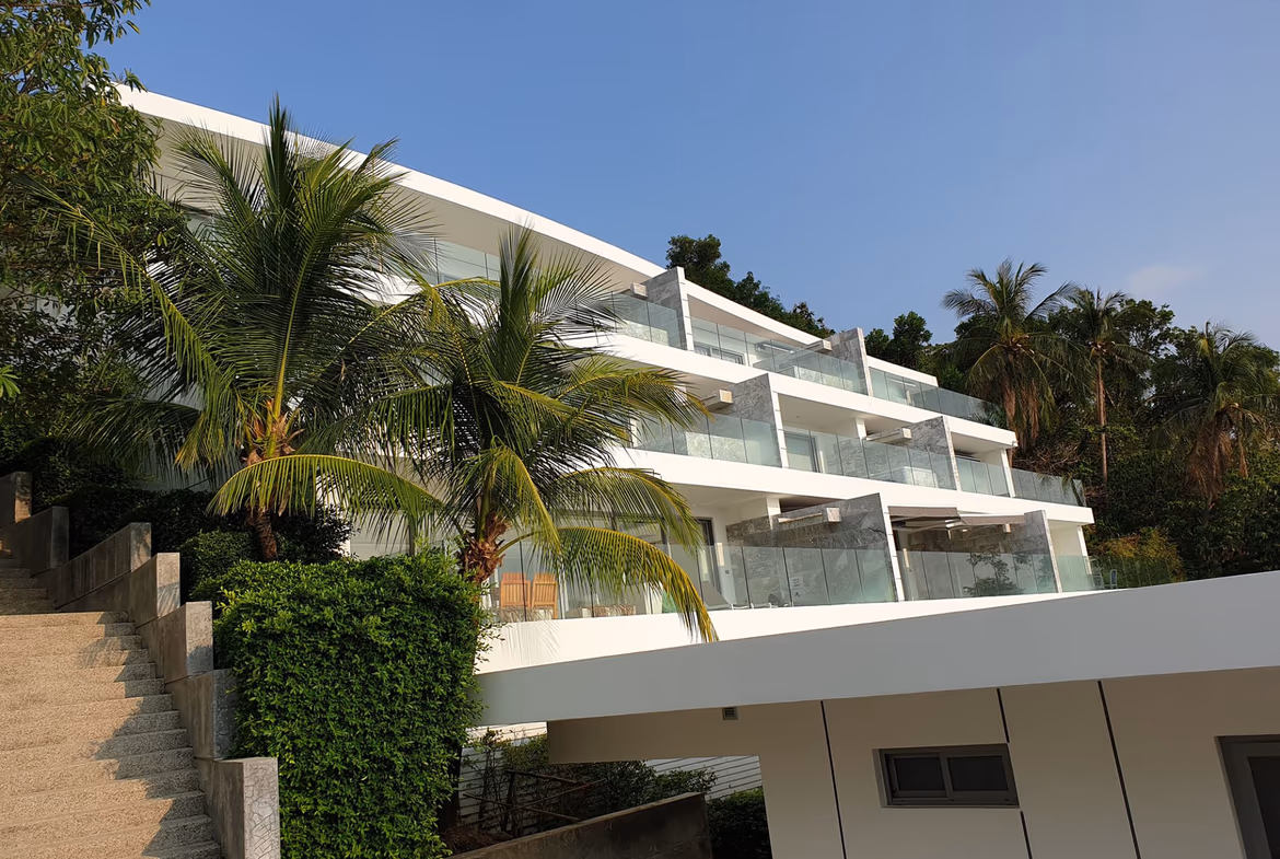 View of a living area with a dark sofa and glass doors leading to a balcony overlooking the sea and distant hills.