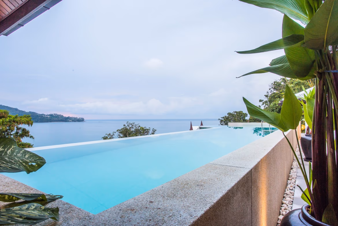 A white 6-bedroom villa overlooks Kamala beach, surrounded by palm trees. The multi-story property features a large infinity pool and several balconies.
