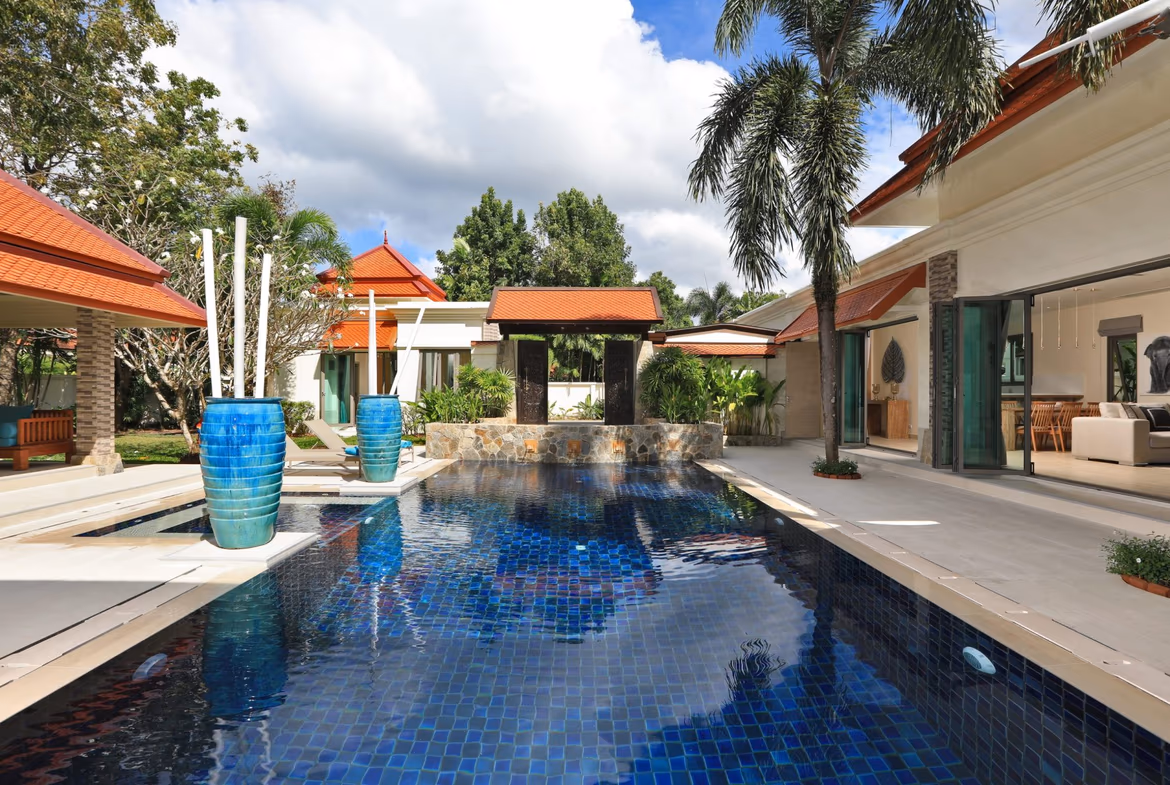 A large, modern 5-bedroom villa in Bang Tao with a flat roof and a swimming pool. The villa's white exterior contrasts with the lush green landscaping.