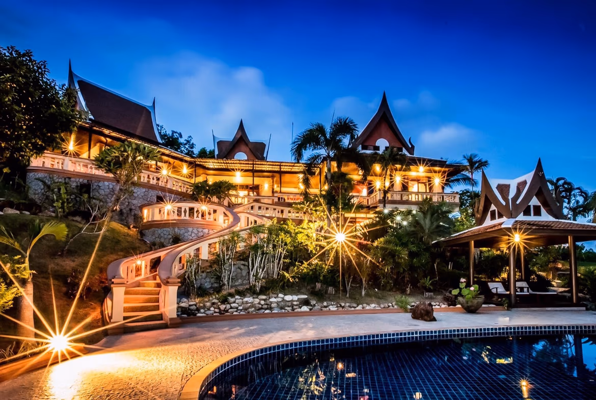 Distant view of a magnificent white villa with a red tile roof, surrounded by palm trees and mountains. This 7-bedroom property in Layan features a large, dark-water swimming pool.