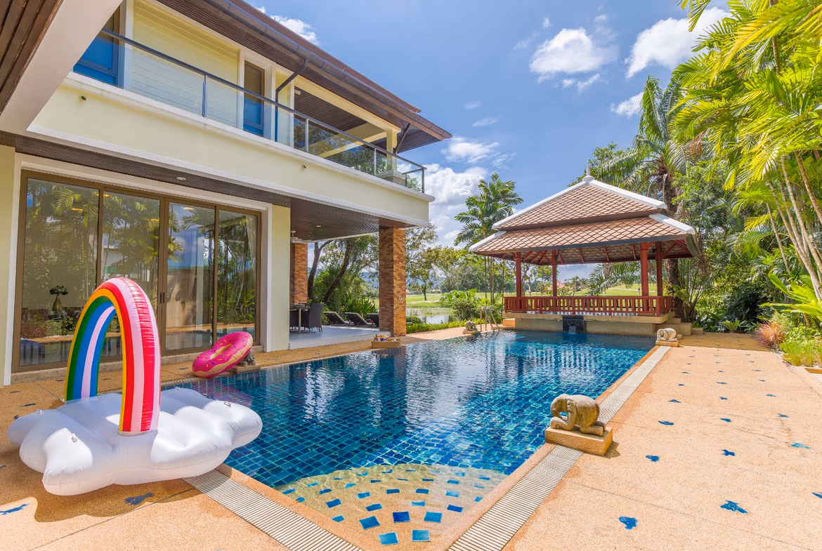 A five-bedroom villa in Phuket features a red tile roof and multiple balconies overlooking a golf course. The expansive home is surrounded by lush green landscaping.