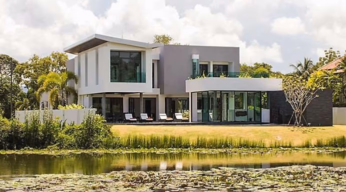 A two-story, 5-bedroom villa in Phuket with a red-tiled roof and an expansive golf course view. The property features a palm-tree-lined driveway leading to the main entrance.
