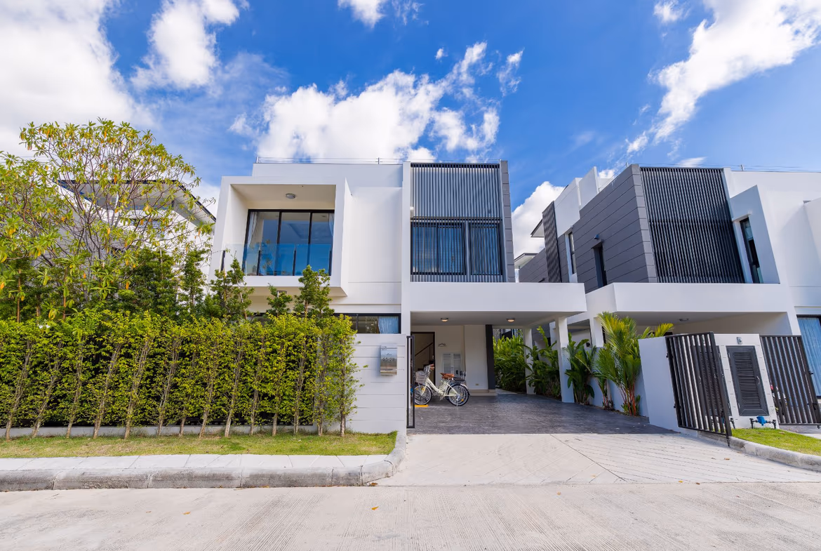 A modern five-bedroom villa in Phuket features a multi-level design and a large infinity pool overlooking a tropical landscape. The white stucco exterior contrasts with the dark-framed windows and wood accents.