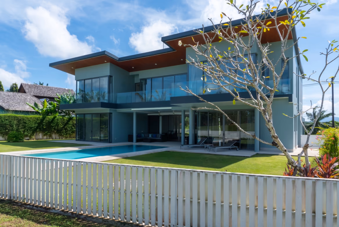 A modern 4-bedroom villa in Phuket features a private infinity pool overlooking the ocean. The white, multi-story villa has large windows and balconies, and is surrounded by lush tropical vegetation.