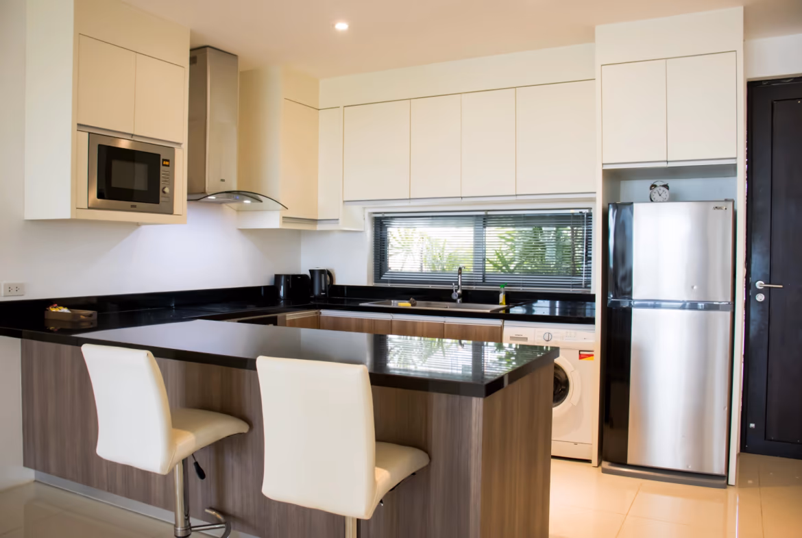 A modern kitchen with light wood cabinetry and a breakfast bar overlooks a living area with a partial sea view.