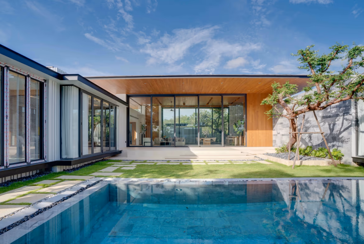 A modern, four-bedroom villa in Bang Tao with a private swimming pool visible in the foreground. The villa features a flat roofline and sits on a body of water under a clear sky.