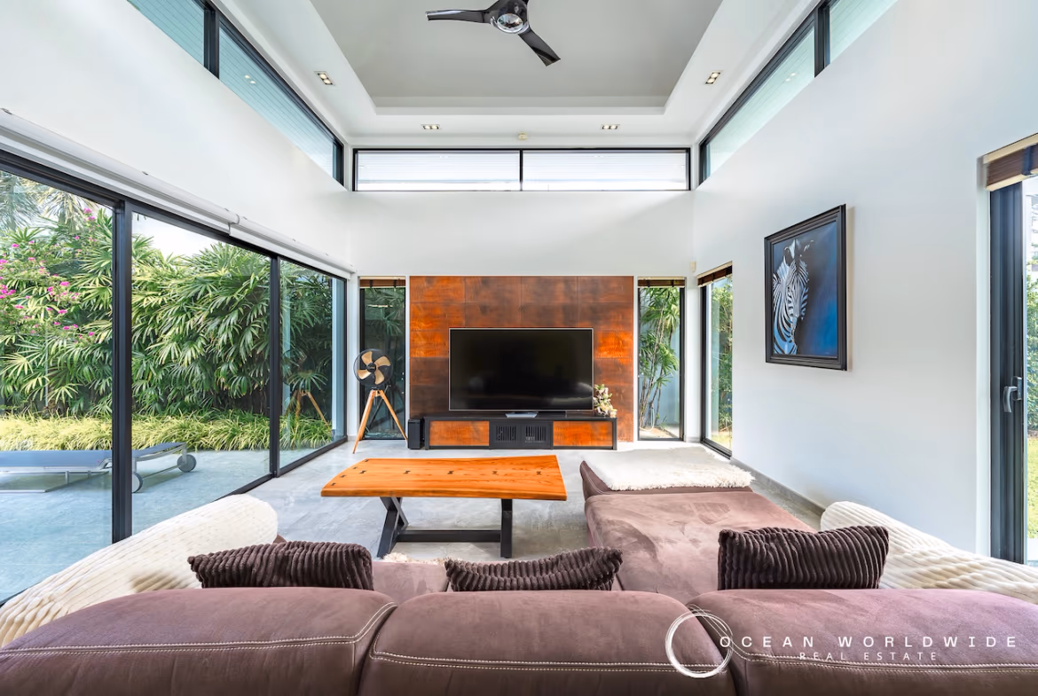 A modern living room with a neutral sofa, patterned rug, and large windows looking out to a garden and pool.