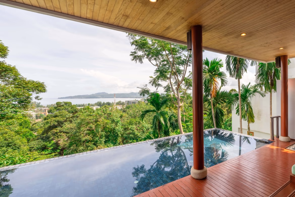 An elevated, head-on exterior view of a four-bedroom villa in Surin, featuring a private swimming pool and a tile roof. The villa has multiple levels and overlooks a lush, treed landscape.