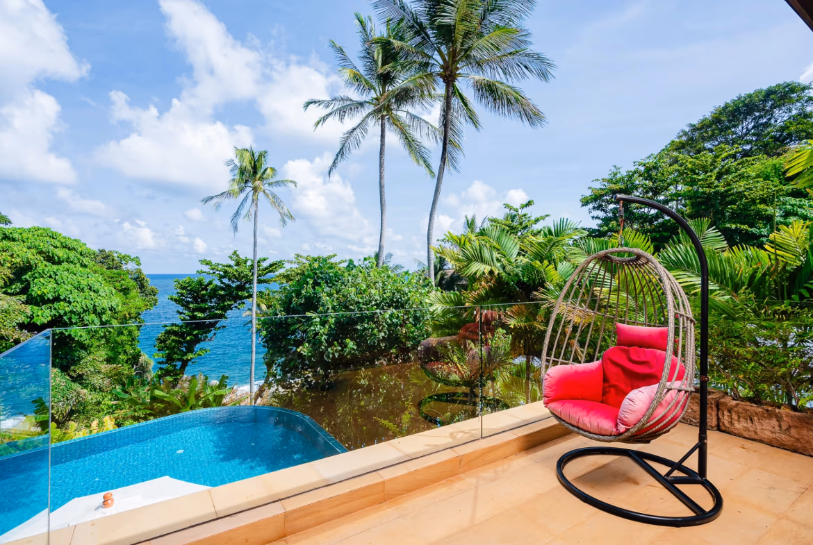 A circular outdoor dining table with four chairs sits on a patio next to a swimming pool, overlooking a tropical landscape and distant sea.
