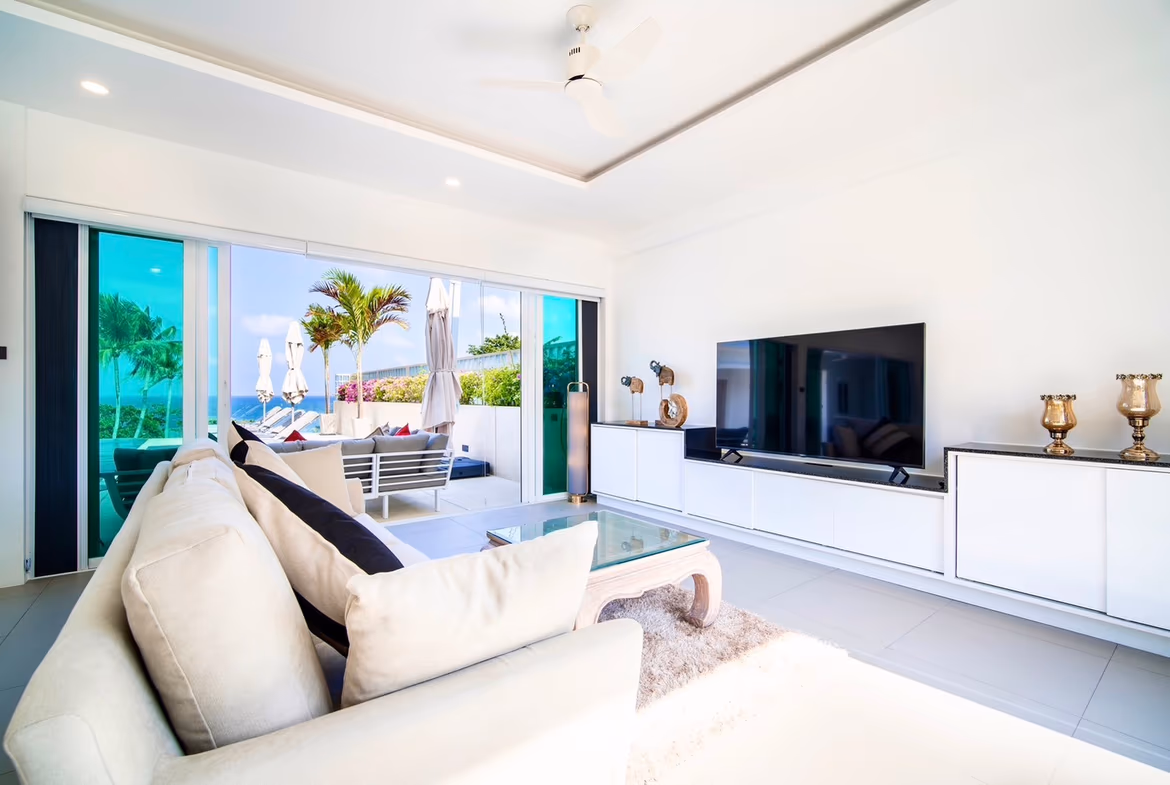A modern living room with a large sofa and television, featuring glass doors that open to a balcony with sea views.