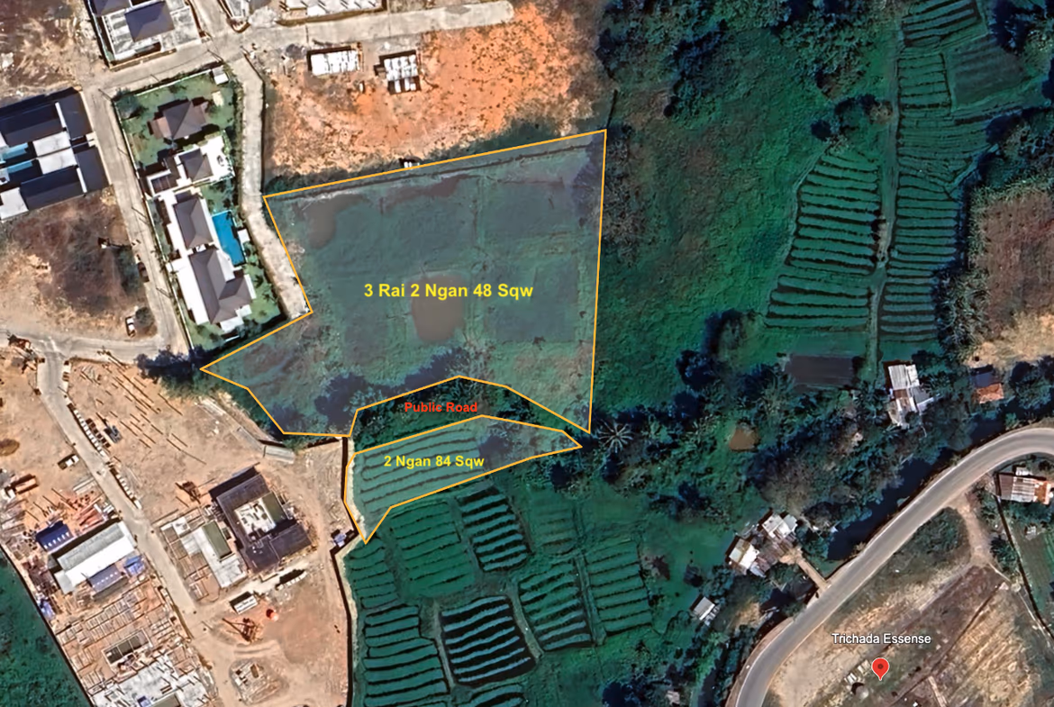 An aerial view shows a large plot of undeveloped land with trees and green foliage, surrounded by other vacant land and some distant buildings.