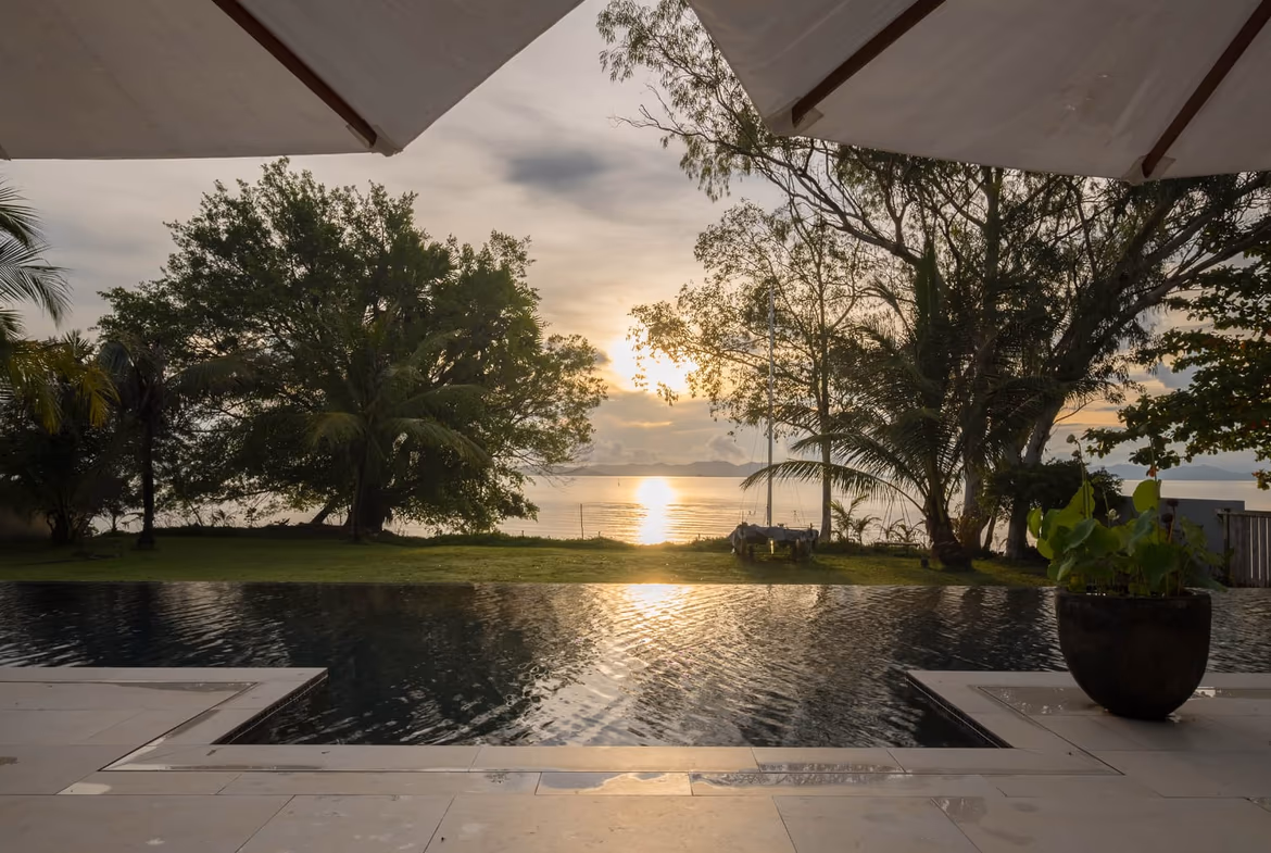 A large, expansive living space featuring a high ceiling, dark wooden beams, light-colored tile floors, and a distant view of the ocean.