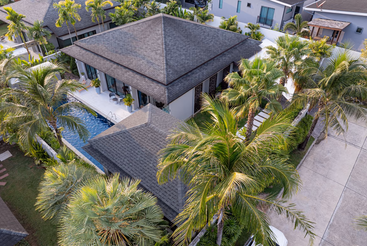 A four-bedroom villa in Phuket features a private pool and a lush, tropical setting. The property's exterior is captured from an elevated angle, showcasing its design and surrounding greenery.