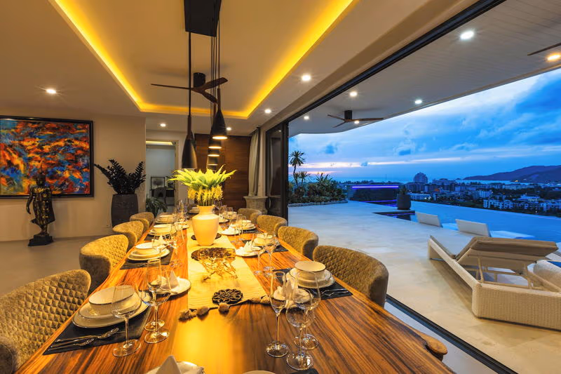 This is a large, six-bedroom villa in Phuket with a private pool overlooking the ocean. The multi-story white building has a large balcony with glass railings, surrounded by palm trees and lush vegetation.