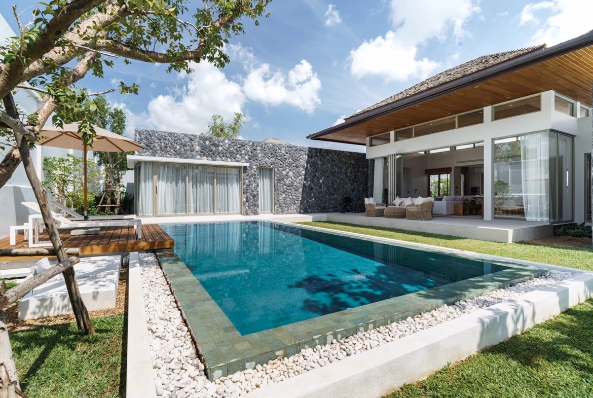 A view of a modern swimming pool with a stone deck and lounge chairs, surrounded by tropical foliage.