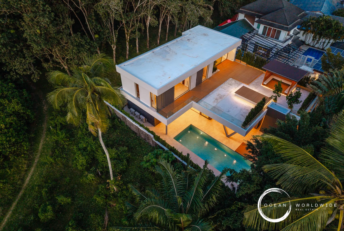 A modern, four-bedroom villa in Layan features a private swimming pool visible from the exterior. The white concrete house has a flat roof and large windows, surrounded by tropical landscaping.