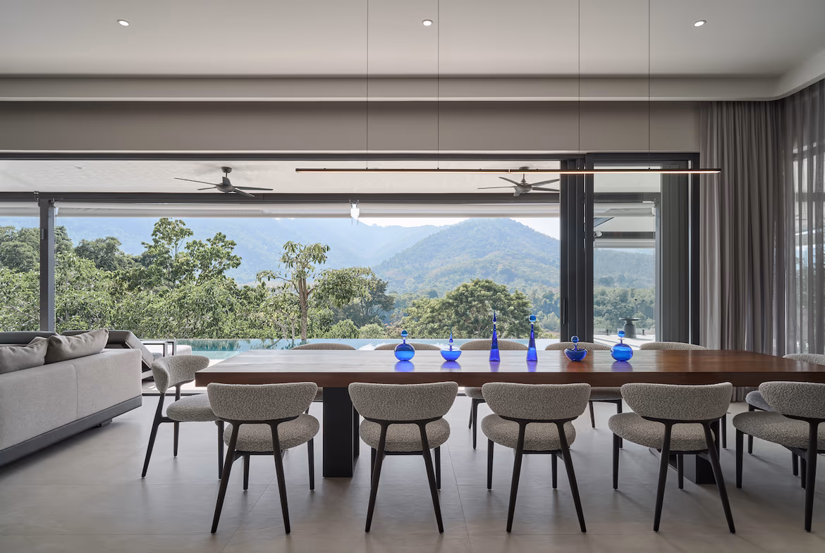A spacious, minimalist living room with white walls and a large, dark framed window overlooks mountains and houses.