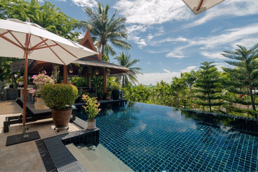 A tropical garden with several palm trees in front of a curved infinity pool overlooking the ocean.