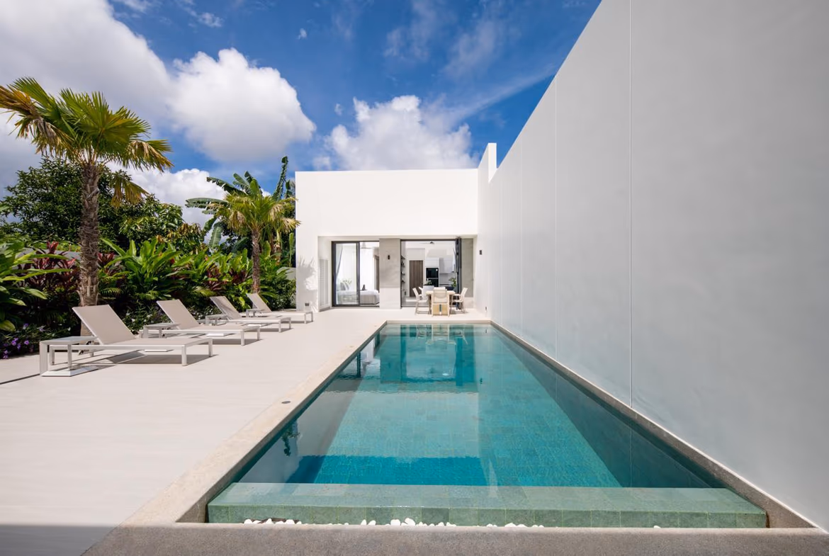 Modern white villa with a long rectangular pool, lounge chairs, and tropical plants under a bright blue sky.