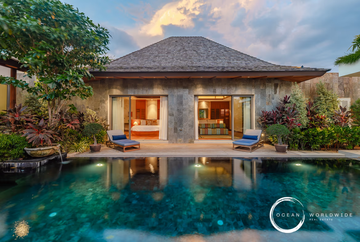 Tropical villa with a stone facade, open glass doors to bedrooms/living area, lush plants, and a reflecting pool in front.