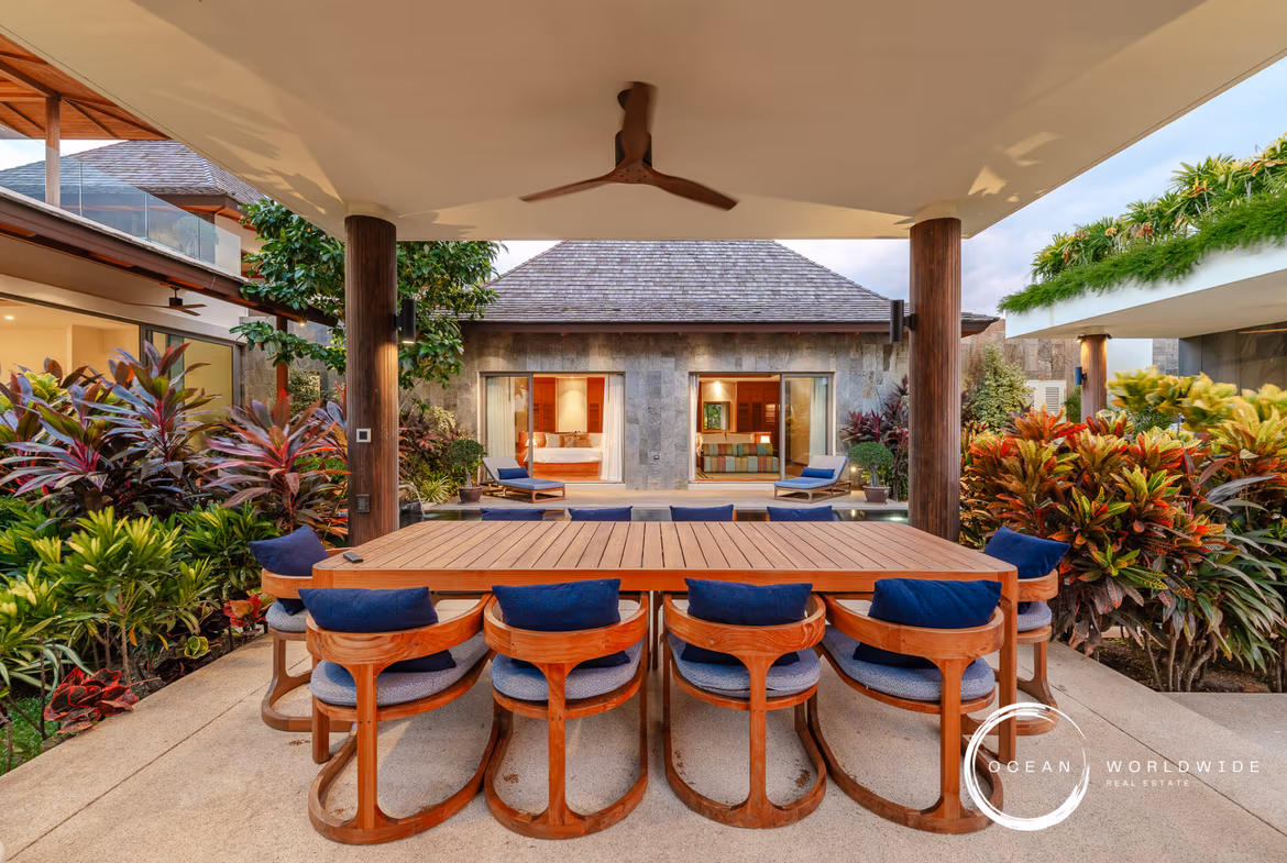 Outdoor dining area with a long wooden table and curved blue-cushioned chairs, surrounded by tropical plants in a villa courtyard under a covered patio.