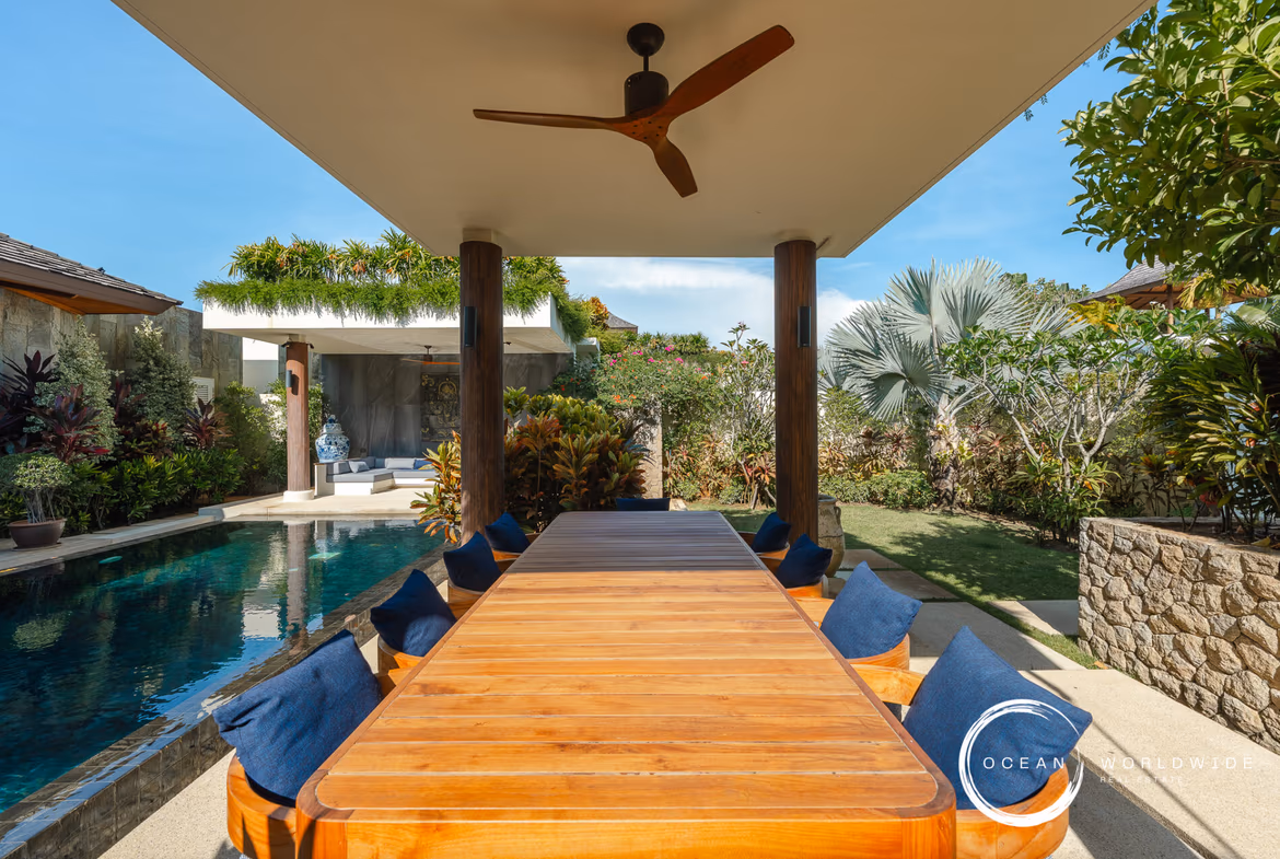 Long wooden outdoor dining table with blue cushions, under a covered patio next to a pool and lush tropical plants.