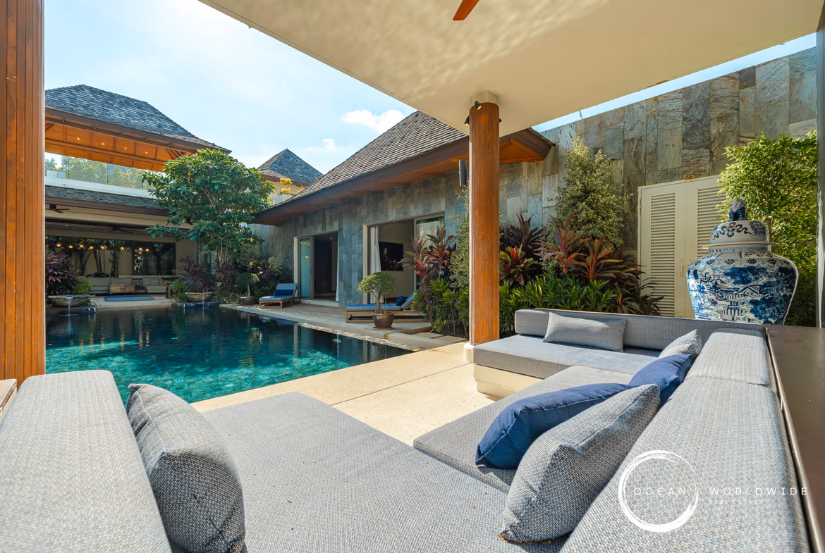 Poolside courtyard of a modern villa with a stone wall, wooden columns, and gray sectional loungers overlooking a turquoise pool.