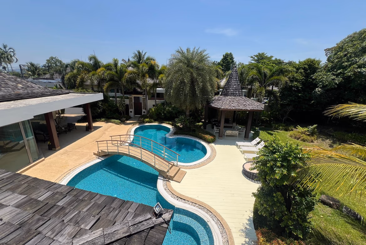 Overhead view of a tropical backyard with a curved blue pool, beige deck, and a thatched gazebo among palm trees.