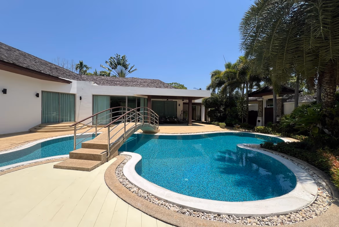 Outdoor backyard with a curved pool, tile steps, and a beige deck beside a white building under a blue sky. Palm trees frame the scene.