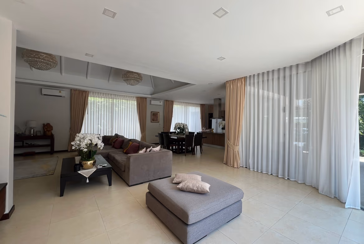 Spacious, modern living room with gray sectional sofa, glass coffee table, and white tiled floor; large windows with sheer curtains let in natural light.