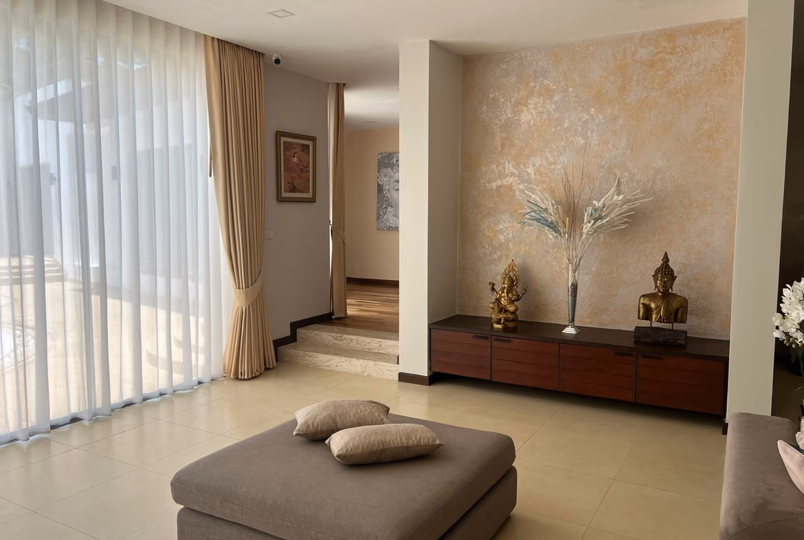 Living room interior with sheer white curtains, beige drapes, and a dark wood console displaying Buddha statues and a tall vase of dried grasses on a textured wall.
