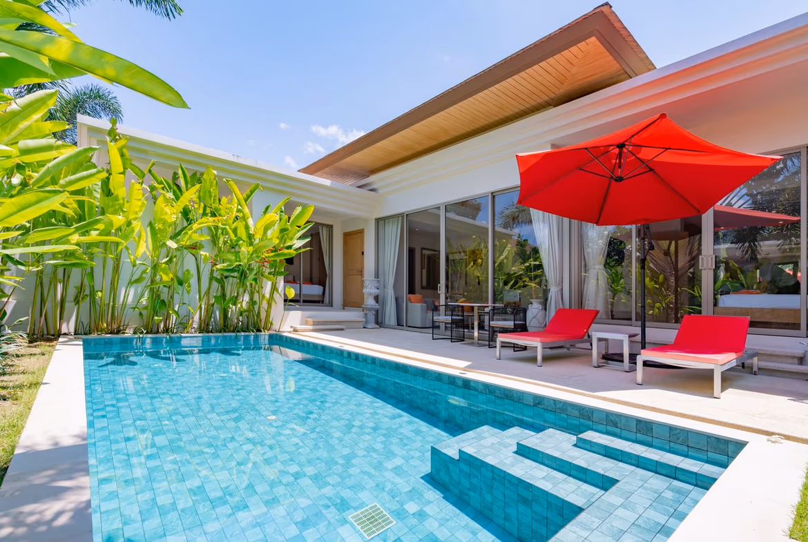 Modern villa poolside: turquoise-tiled pool, red lounge chairs, and a large red patio umbrella by glass doors. A tropical garden lines the left side.