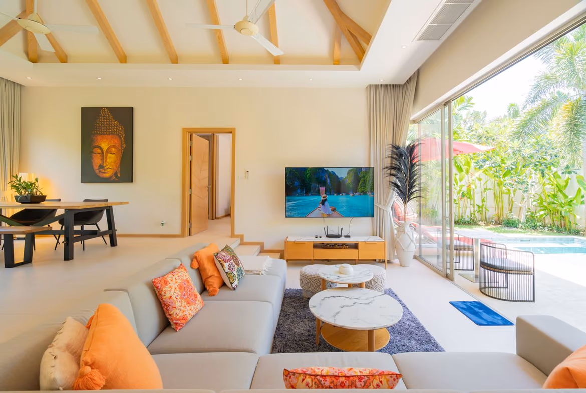 Bright living room with a beige sectional, orange cushions, and marble coffee tables facing a TV; glass doors overlook a pool and tropical garden.
