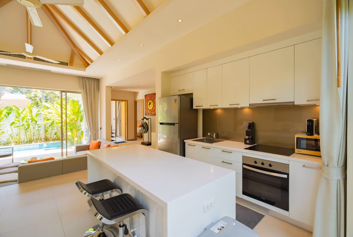 Open-plan kitchen with white island, bar stools, and stainless steel appliances adjacent to a living area and sliding glass doors to a lush outdoor pool area.