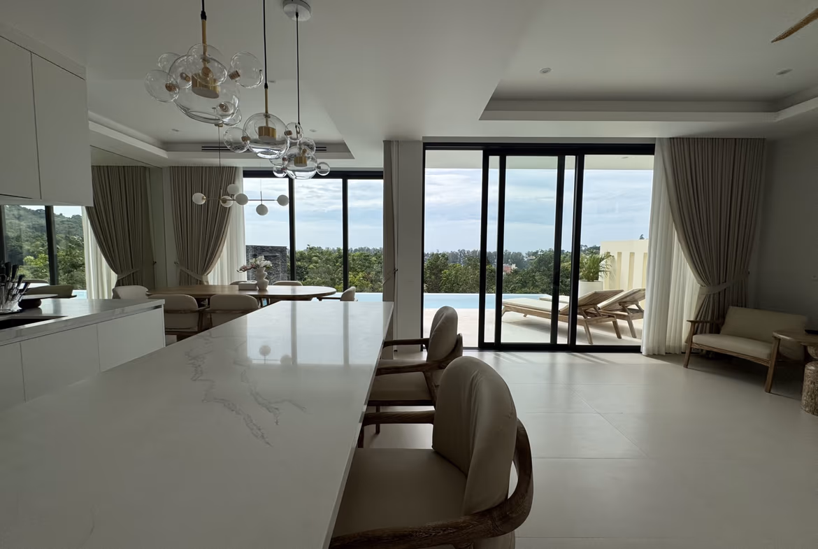 Open-plan kitchen and dining area with a white marble island, wicker chairs, and large glass doors opening to a pool and outdoor seating.