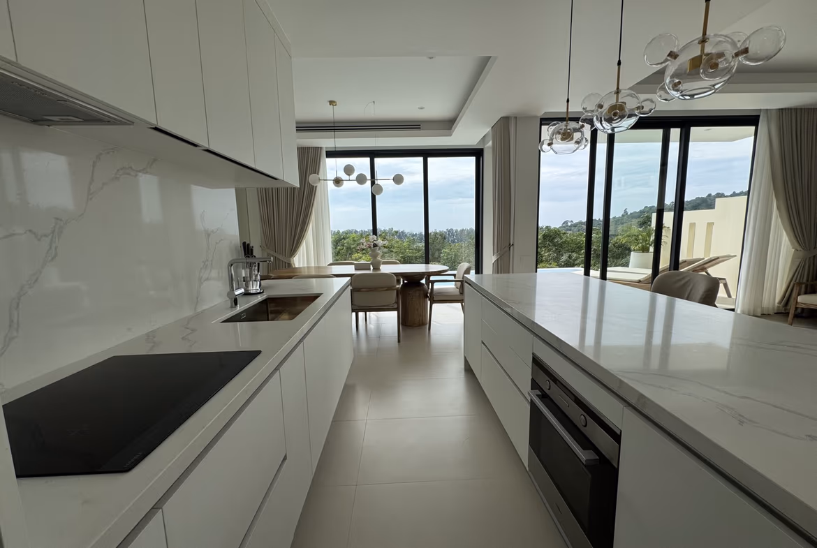 Contemporary white kitchen with marble countertops, an island, and built-in oven; large windows overlook a greenery landscape.