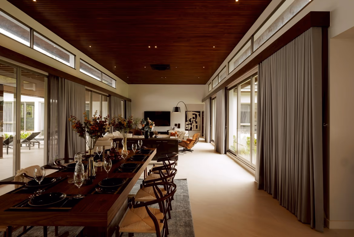 Spacious dining room with a long wooden table set for a formal meal, surrounded by glass walls and a dark wood ceiling.