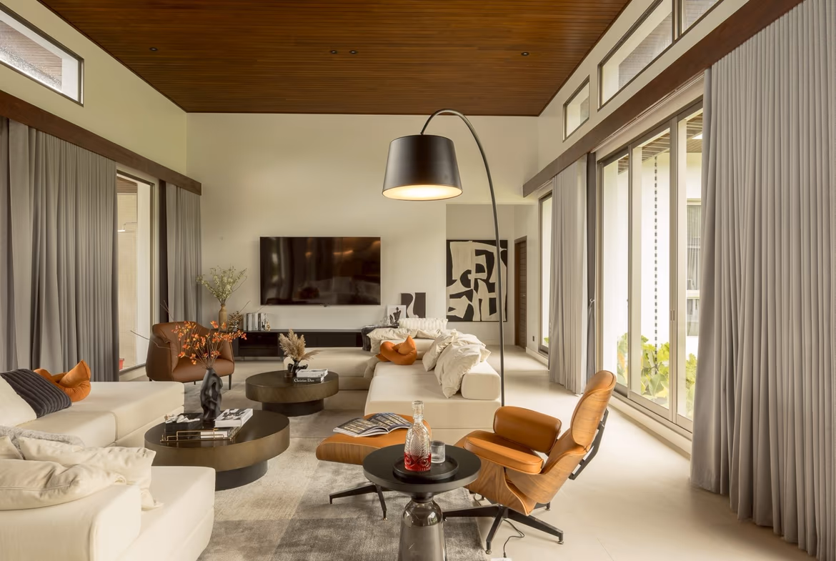 Modern living room with white sofas, orange chairs, and round black coffee tables under a wood-paneled ceiling. A curved arc floor lamp stands center stage.