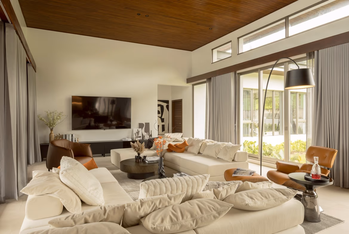 Spacious living room with a cream sectional, orange pillows, and a dark coffee table, near large windows and a TV wall.